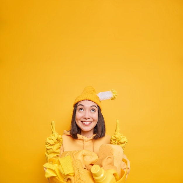 空白の上のアジアの女性用務員ポイントは、ランドリー バスケットに掃除用品を運び、家事に最適な製品を示し、黄色い壁に帽子のゴム手袋をはめている