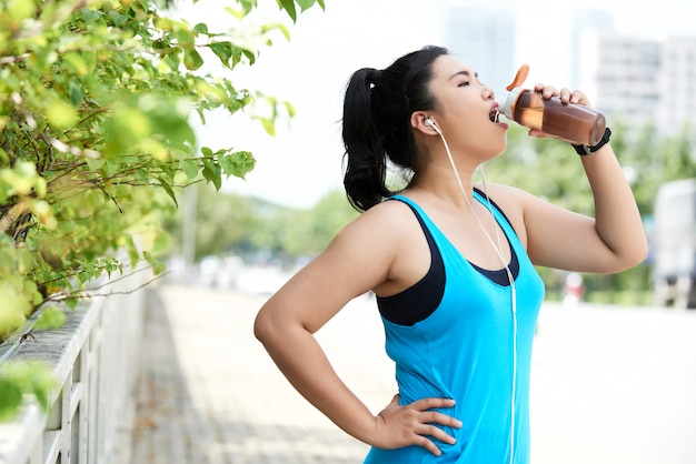 Asian female jogger drinking energy shake from sports bottle in street
