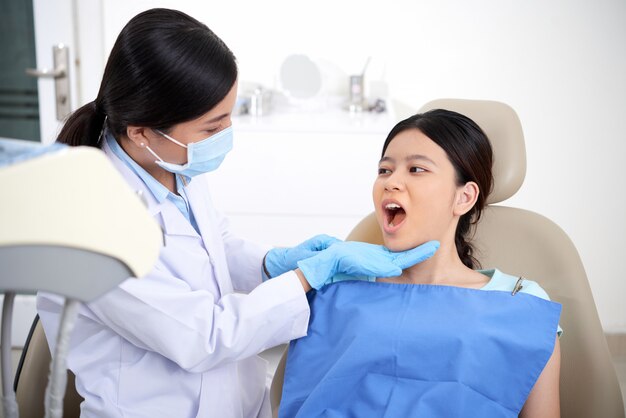 Азиатский женский пациент, сидя в кресле с открытым ртом, и стоматолог, глядя на ее зубы
