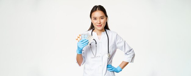 Asian female physician showing pills medication holding vitamins and wearing rubber gloves standing over white background