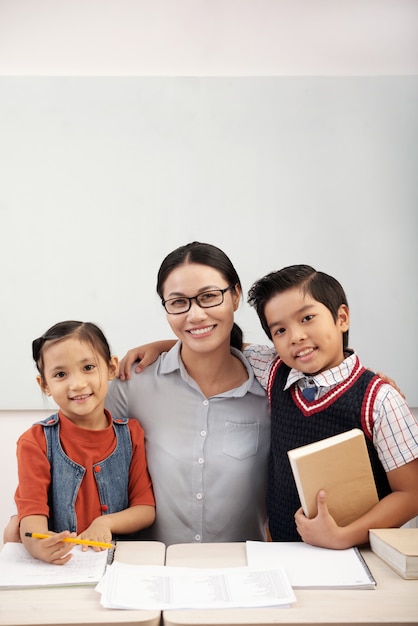 Asian female teacher in glasses posing in classroom with boy and girl pupils