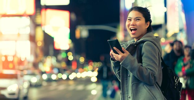 アジアの女性旅行者の笑顔の幸せの手持ちミラーレスカメラは、冬の季節に明るいボケ味と人々の群衆が背景をぼかすナイトライフライフスタイルのトーロストをお楽しみください