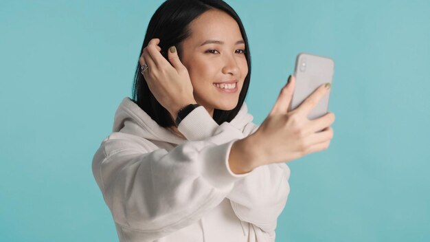 Asian female wearing hoodie preening and taking selfie on smartphone over colorful background Modern technology Beautiful woman posing in studio