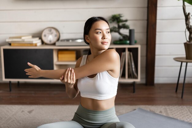 Asian fitness girl doing workout at home sitting on floor mat in activewear warm up and stretching a...