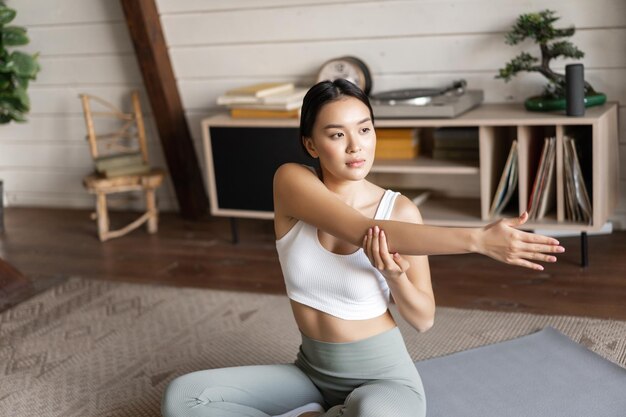 Азиатская фитнес-девушка тренируется дома, сидя на коврике в спортивной одежде, разогреваясь и растягивая ...