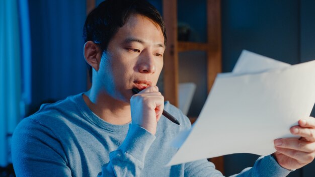 Asian freelance businessman focus work type on laptop computer busy with full of paperwork chart on desk in living room