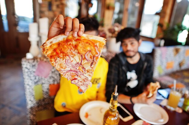 피자 가게에서 파티 중 피자를 먹는 아시아 친구들 행복한 인도 사람들이 함께 이탈리아 음식을 먹고 소파에 앉아 즐거운 시간을 보내고 있습니다.