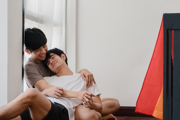 Asian Gay couple lying and hugging on the floor at home. Young Asian LGBTQ+ men kissing happy relax rest together spend romantic time in living room with rainbow flag at modern house in the morning.