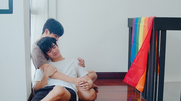 Asian gay couple lying and hugging on the floor at home. young asian lgbtq+ men kissing happy relax rest together spend romantic time in living room with rainbow flag at modern house in the morning.