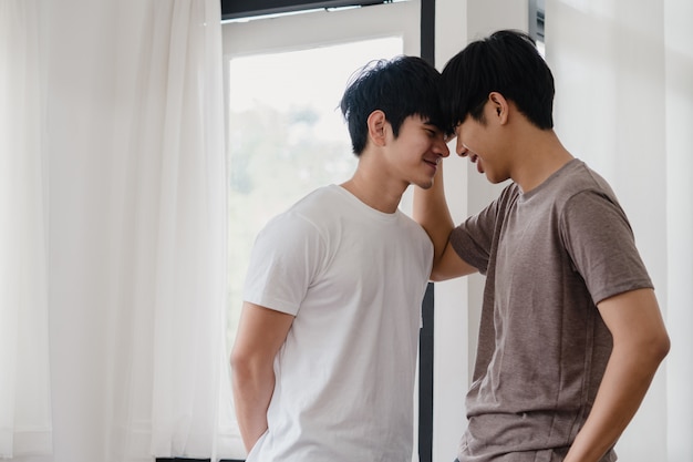 아시아 게이 커플 서 집에서 창문 가까이 포옹. 행복 키스 젊은 아시아 LGBTQ + 남자는 아침에 현대 집에서 거실에서 낭만적 인 시간을 보내고 휴식을 취하십시오.
