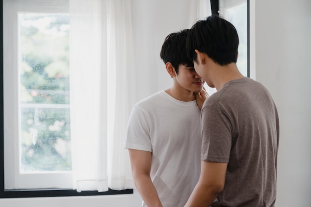 아시아 게이 커플 서 집에서 창문 가까이 포옹. 행복 키스 젊은 아시아 LGBTQ + 남자는 아침에 현대 집에서 거실에서 낭만적 인 시간을 보내고 휴식을 취하십시오.