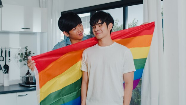 Asian Gay couple standing and hugging room at home. Young handsome LGBTQ+ men kissing happy relax rest together spend romantic time in modern kitchen with rainbow flag at house in the morning .