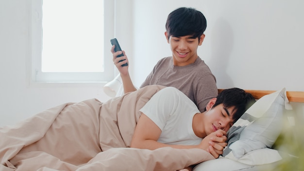 Asian Gay couple using mobile phone at home. Young Asia LGBTQ+ man happy relax rest after wake up, check social media while his boyfriend sleep lying on bed in bedroom at home in the morning .