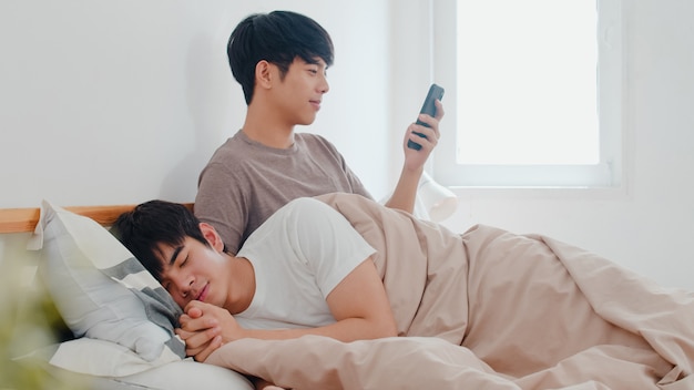 Asian Gay couple using mobile phone at home. Young Asia LGBTQ+ man happy relax rest after wake up, check social media while his boyfriend sleep lying on bed in bedroom at home in the morning .