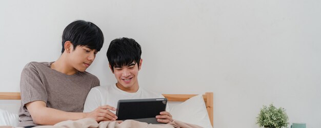 Азиатские пары гомосексуалиста используя таблетку дома. Молодые азиатские LGBTQ + мужчины счастливы расслабиться вместе отдохнуть после пробуждения, проверки почты и социальных сетей, лежа на кровати в спальне дома утром.