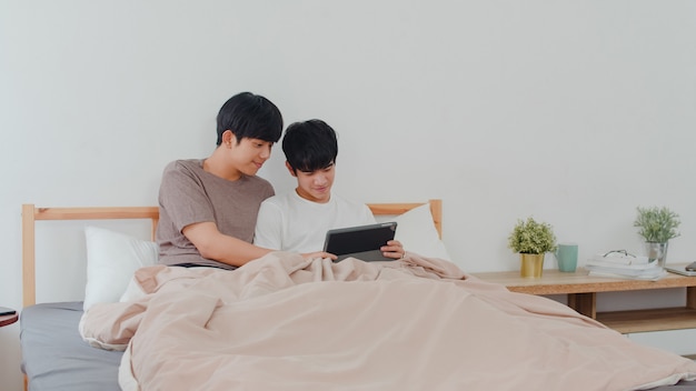 Азиатские пары гомосексуалиста используя таблетку дома. Молодые азиатские LGBTQ + мужчины счастливы расслабиться вместе отдохнуть после пробуждения, проверки почты и социальных сетей, лежа на кровати в спальне дома утром.