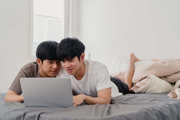 Asian Gay lgbtq men couple using computer laptop at modern home. Young Asia lover male happy relax rest together after wake up, watching movie lying on bed in bedroom at house in the morning .