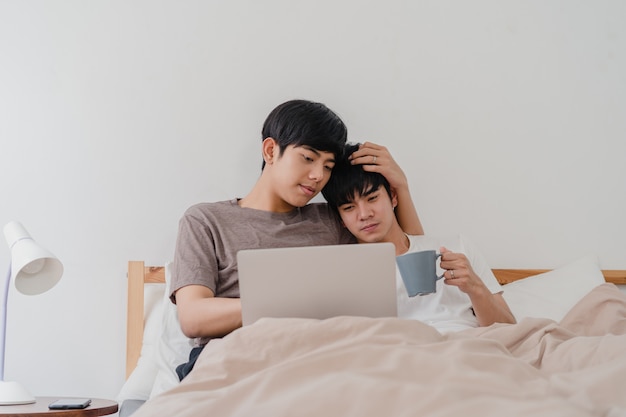 Asian Gay men couple using computer laptop and drinking coffee at modern home. Young Asia lover male happy relax rest together after wake up, watching movie lying on bed in bedroom at house in morning