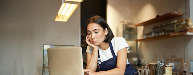 무료 사진 카페에서 카운터 뒤에 서있는 아시아 소녀 바리스타는 느낌이있는 동안 노트북으로 일하고 있습니다.