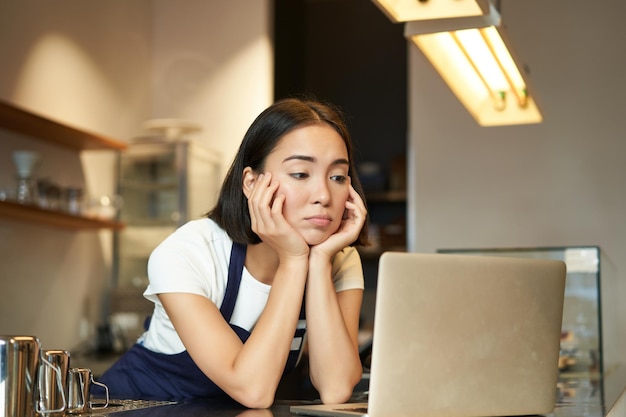 カフェのカウンターの後ろに立っているアジアの女の子のバリスタは、憂鬱を感じながらノートパソコンを見て