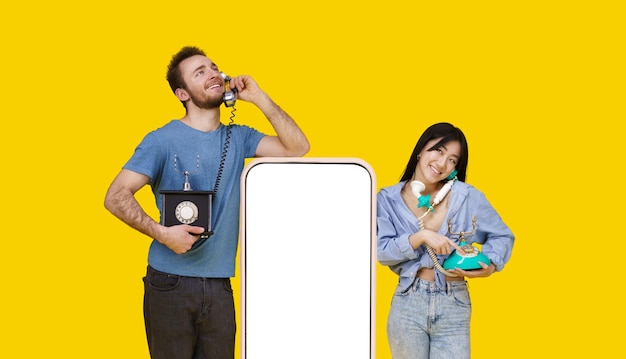 Asian girl and caucasian guy in love talking on vintage phones leaned on huge smartphone with blank screen