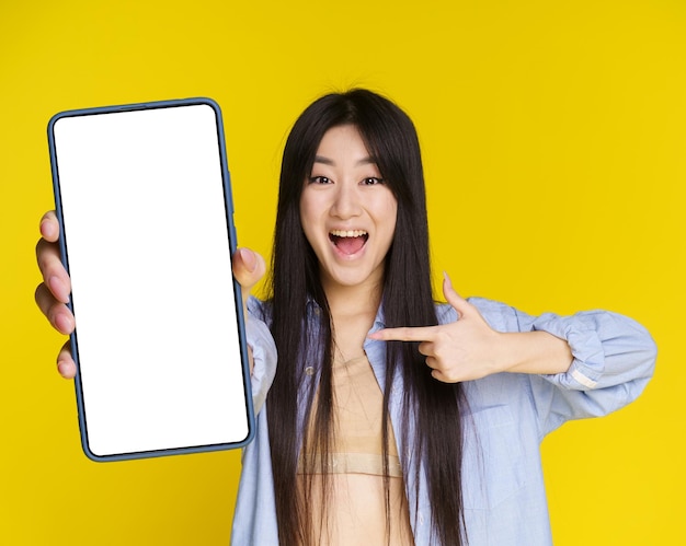 アジアの女の子は、黄色の背景で隔離された空の画面を指しているスマートフォンを保持している宝くじカジノゲームの販売オファーに興奮しています製品の配置は、モバイルアプリケーションの広告をモックアップします