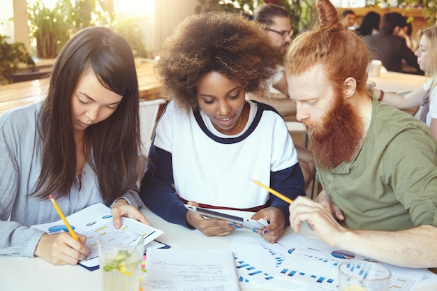 Asian girl filling in papers with graphs and diagrams while African woman sharing ideas with redhead bearded colleague on touch pad.