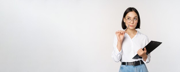 Asian girl in glasses thinks holds pen and clipboard writing down making notes standing over white background