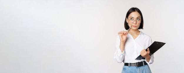 Азиатская девушка в очках думает, что держит ручку и буфер обмена и записывает, делая заметки, стоя на белом фоне