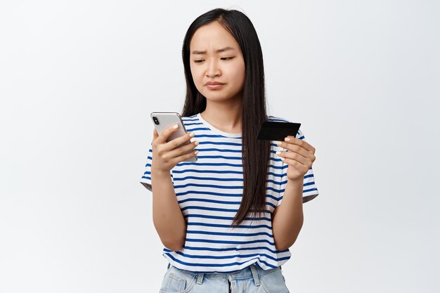 Азиатская девушка испытывает трудности с проблемой приложения для покупок в Интернете во время оплаты, хмурясь на экране мобильного телефона, держа кредитную карту, стоящую на белом фоне