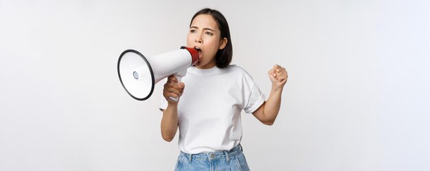 Asian girl shouting at megaphone young activist protesting using loud speakerphone standing over white background Copy space