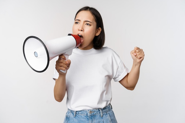 Asian girl shouting at megaphone young activist protesting using loud speakerphone standing over white background