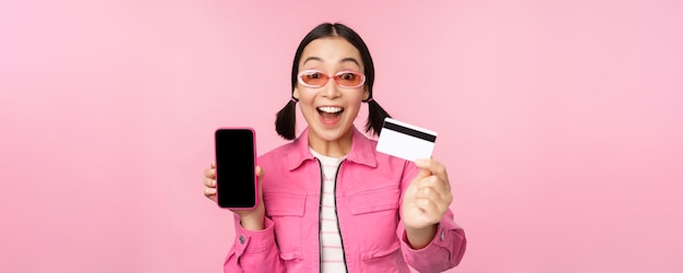 Азиатская девушка показывает экран мобильного телефона и реагирует на кредитную карту, удивленная камерой, задыхаясь от впечатлений