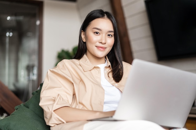 Asian girl sitting in living room with laptop woman watching videos on computer at home relaxing wit...