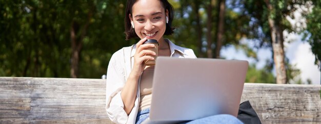 아시아 소녀 가 노트북 과 무선 이어폰 을 들고 앉아 커피 를 마시고 스크린 을 보고 있다