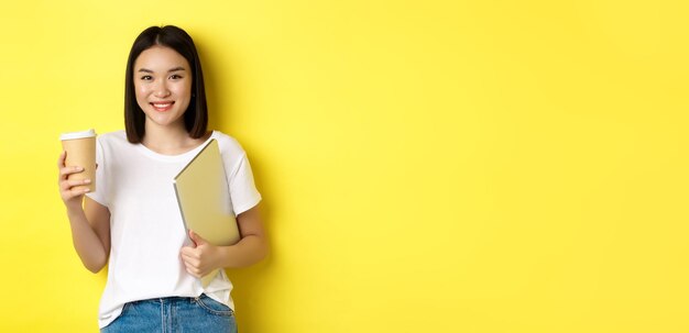 Asian girl student drinking coffee and holding laptop smiling at camera standing over yellow backgro