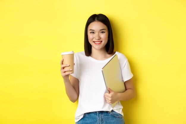 노란색 배경 위에 서서 커피를 마시고 노트북을 들고 카메라를 보며 웃고 있는 아시아 여학생