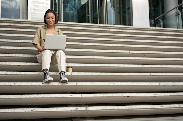 Foto gratuita la studentessa asiatica si siede sulle scale vicino ai tipi del campus sul computer portatile fa i compiti all'aperto