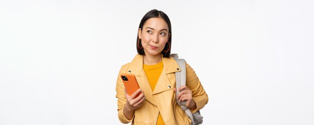 Asian girl traveller with backpack holding mobile phone using smartphone app looking thoughtful standing over white background