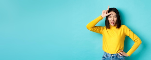 Free photo asian girl winner mocking over lost team showing loser sign on forehead and sticking tongue ignorant