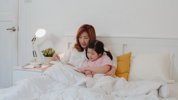 Asian grandmother read fairy tales to granddaughter at home. Senior Chinese, grandma happy relax with young girl enjoy good quality time lying on bed in bedroom at home at night concept.