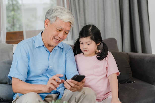 Asian grandparents and granddaughter using mobile phone at home. Senior Chinese, grandpa and kid happy spend family time relax with young girl checking social media, lying on sofa in living room.