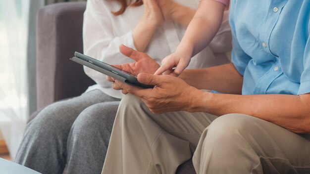 Asian grandparents and granddaughter using tablet at home. Senior Chinese, grandpa and grandma happy spend family time relax with young girl checking social media, lying on sofa in living room concept