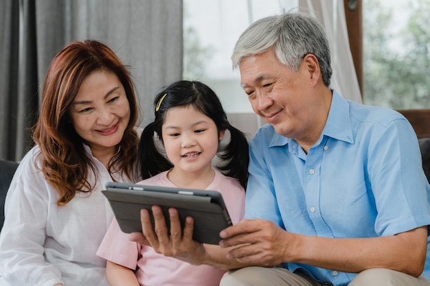 Asian grandparents and granddaughter video call at home. Senior Chinese, grandpa and grandma happy with girl using mobile phone video call talking with dad and mom lying in living room at home.