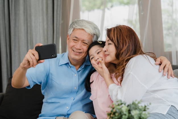 Asian grandparents selfie with granddaughter at home. Senior Chinese, grandpa and grandma happy spend family time relax using mobile phone with young girl kid lying on sofa in living room concept.