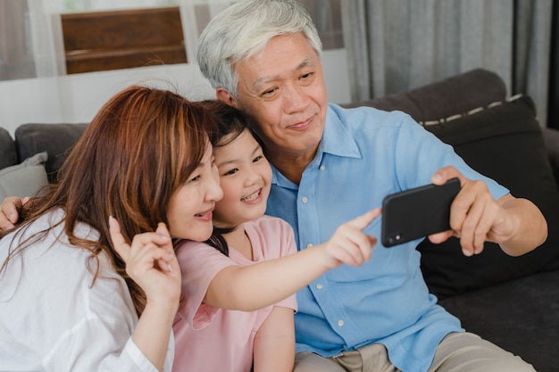 Asian grandparents selfie with granddaughter at home. Senior Chinese, grandpa and grandma happy spend family time relax using mobile phone with young girl kid lying on sofa in living room concept.