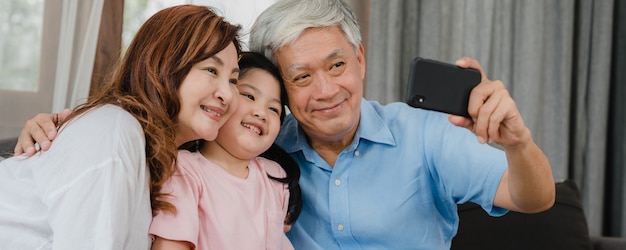 Asian grandparents selfie with granddaughter at home. Senior Chinese happy spend family time relax using mobile phone with young girl kid lying on sofa in living room.