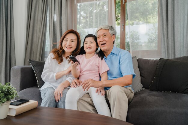 Asian grandparents watch TV with granddaughter at home. Senior Chinese, grandfather and grandmother happy using family time relax with young girl kid lying on sofa in living room concept.