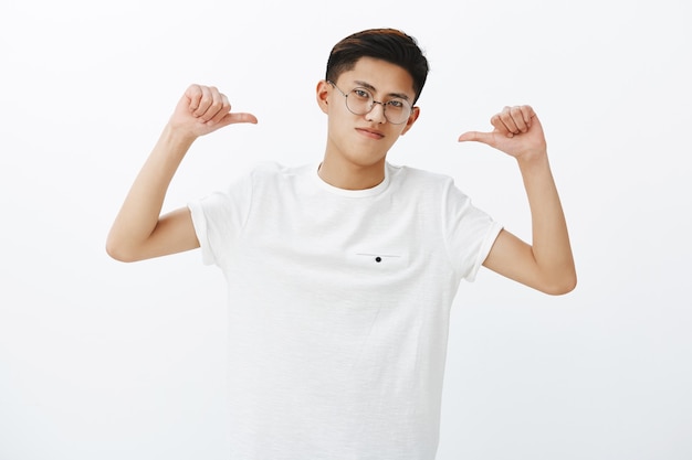 Asian guy is pro feeling proud and self-assured triumphing, being winner pointing at himself with thumbs tilting head looking confident and assertive in own skills