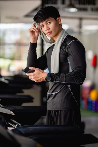 スポーツウェアとスマートウォッチを身に着けているアジアのハンサムな男性は、トレッドミルでタオルを使用して額の汗を拭き、現代のジムでのトレーニング後にスマートフォンを持っています。